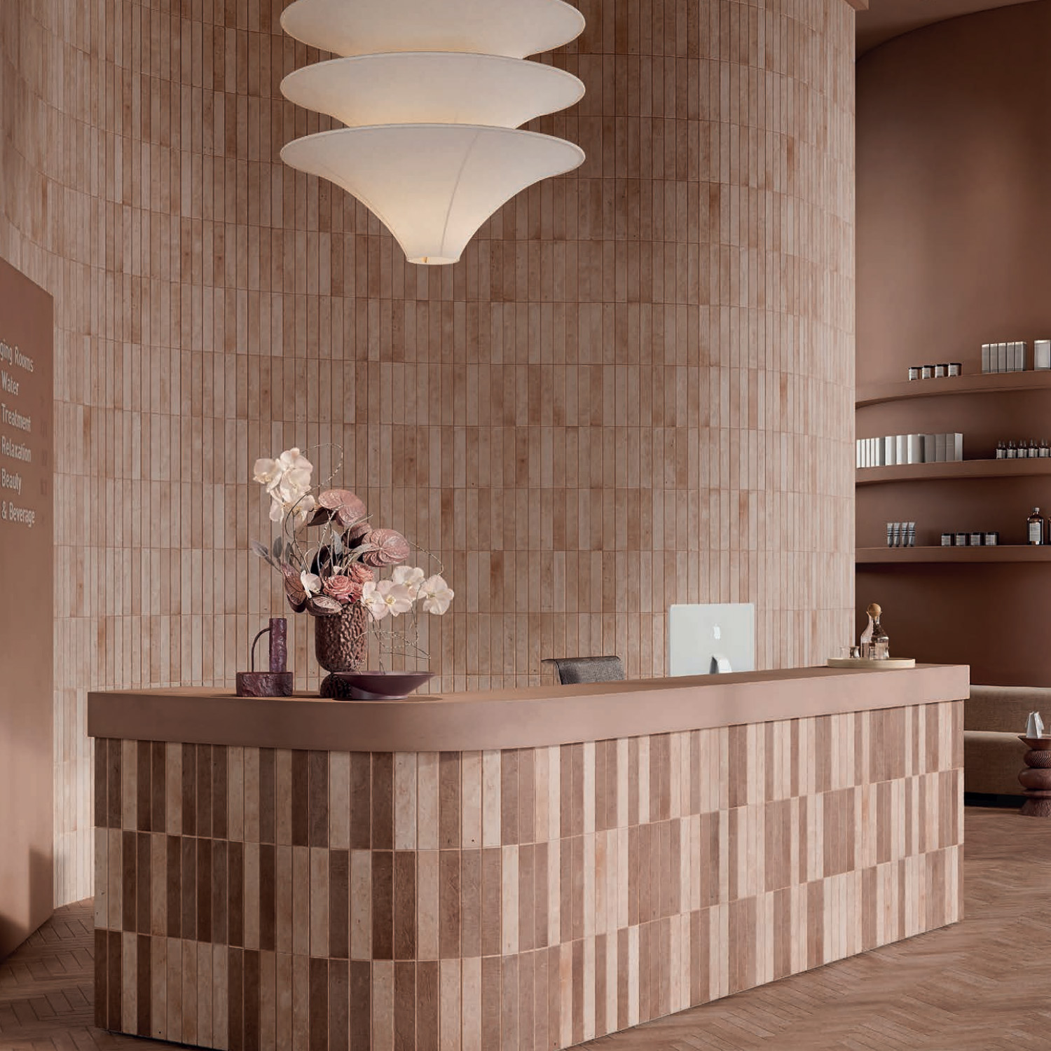 A modern reception area featuring a tan tiled desk, a computer, a vase with pink flowers, neutral-toned walls, shelves with products, and the Tyrrhenia - Mauve large white layered pendant light overhead.