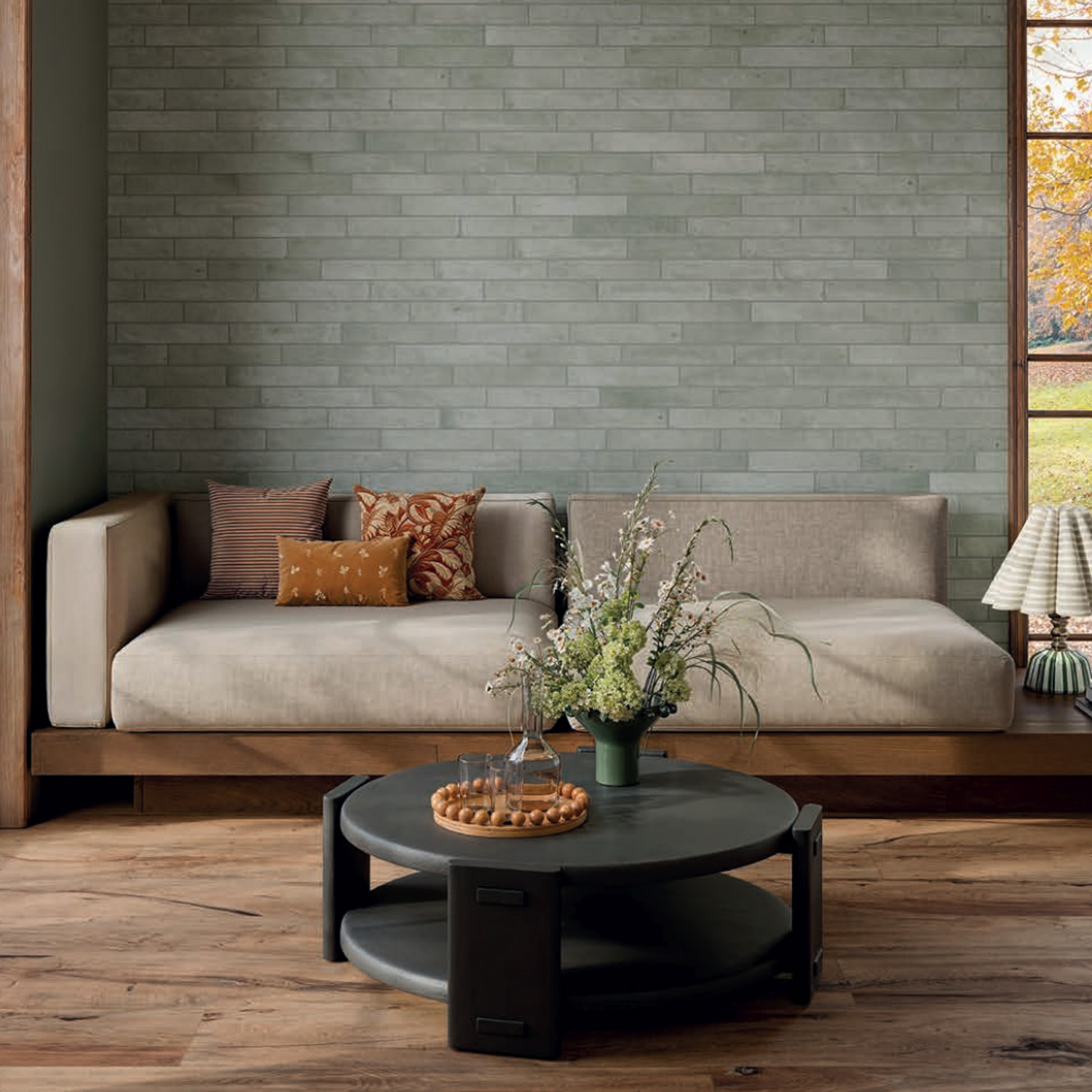 A modern living room featuring the Tyrrhenia - Sage sofa, complemented by assorted cushions, a round black coffee table with a flower vase, and wood flooring. The space is framed by light gray brick walls and large windows showcasing trees and grass outside.