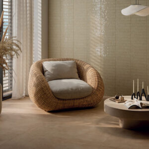 A cozy Via Del Sale - Beige wicker chair with a cushion sits by a window with sheer curtains. A round coffee table holds books, candles, and glasses, while neutral tones and dried flowers create a calm, modern living space.