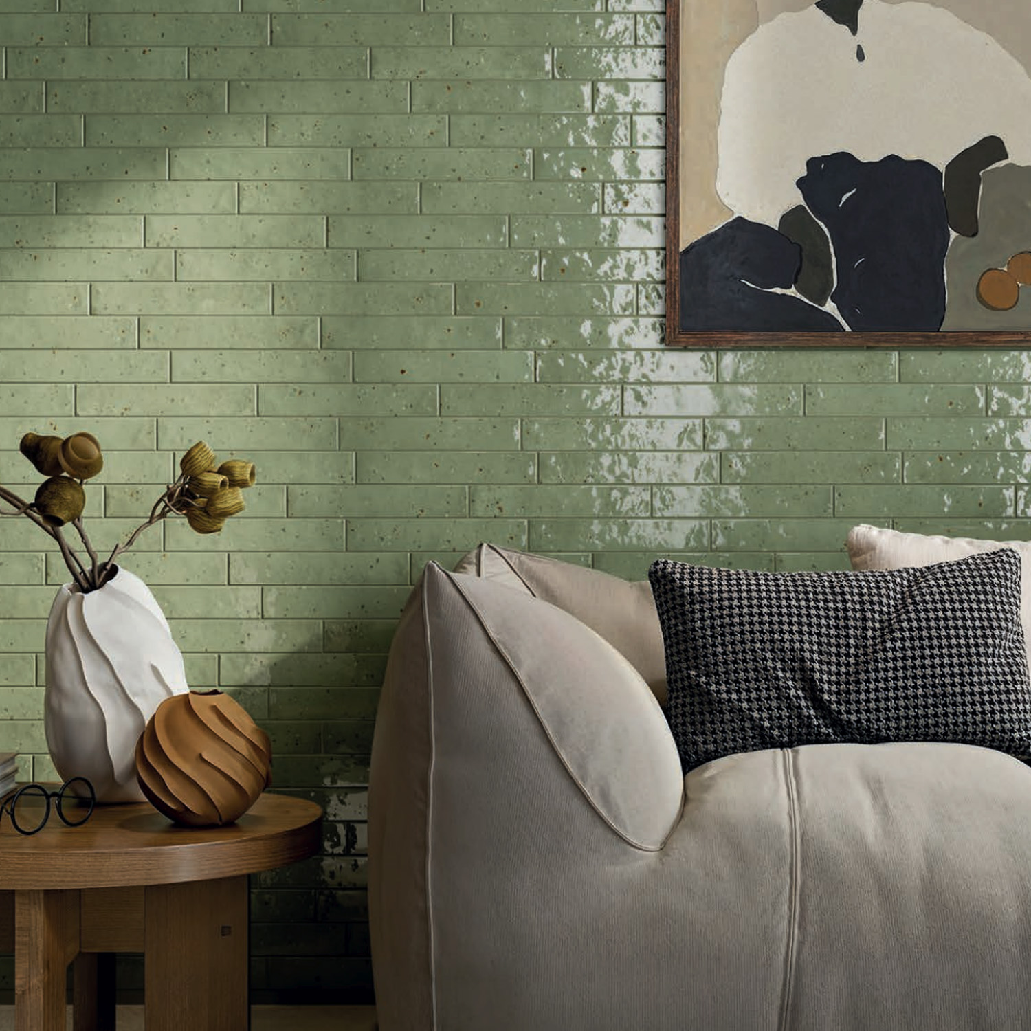 A cozy beige sofa with textured pillows sits beside a wooden table adorned with decorative vases, set against a Via Del Sale - Green tiled wall and an abstract art piece.