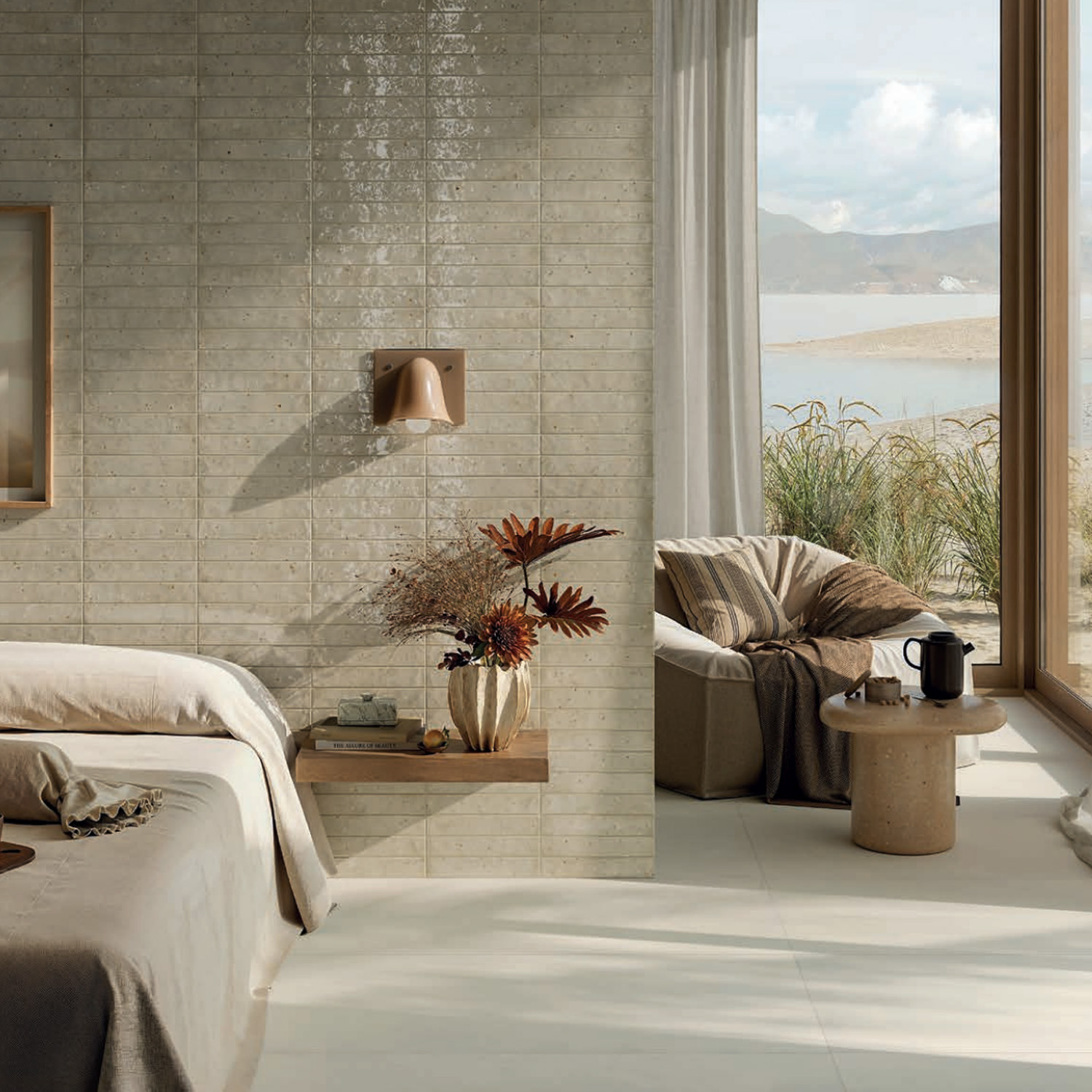 A cozy bedroom featuring the Via Del Sale - Mud bed, a floating nightstand adorned with a vase of dried flowers, a lounge chair, and a small table holding a mug. Large windows offer stunning views of sand dunes and water outside.