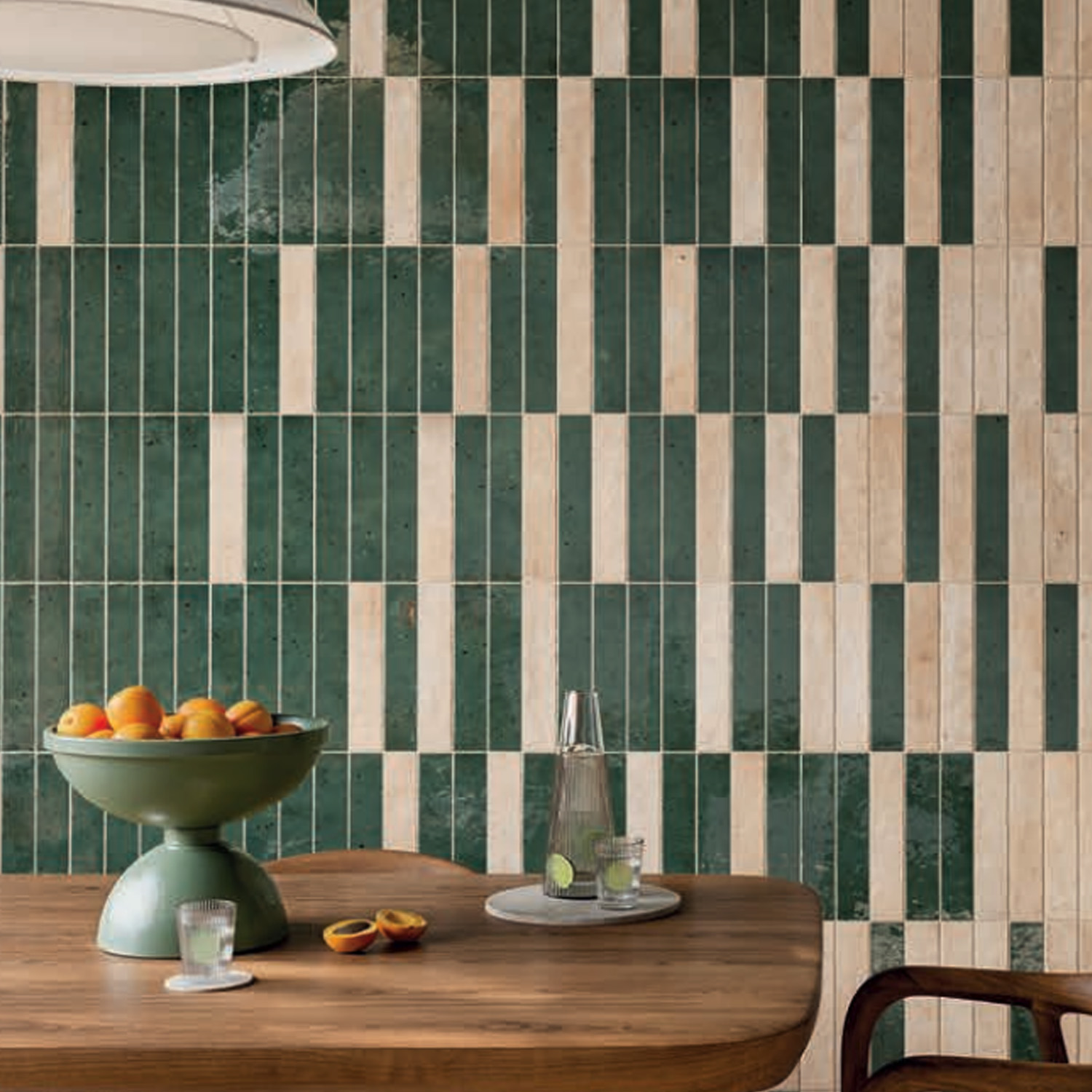 A wooden table features a green fruit bowl filled with oranges and peaches, a glass of water, a glass pitcher with lime slices, and two peach halves. The wall in the background is adorned with Via Del Sale - Rain Forest tiles, arranged vertically in green and beige stripes.