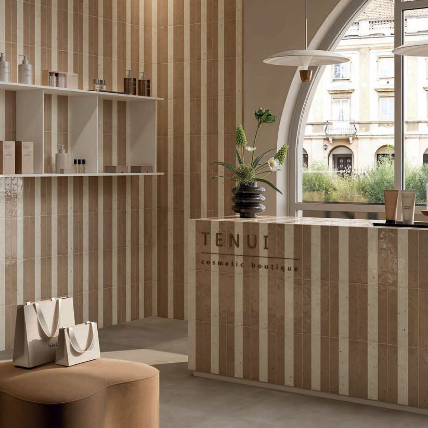 Modern boutique interior featuring Via Del Sale - Sand on the beige striped walls, a reception counter, white shelves displaying products, shopping bags on a pouf, a vase with flowers, and a large arched window overlooking a street with buildings.