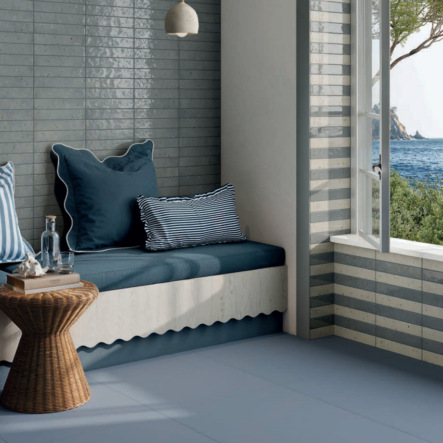 Cozy reading nook with Via Del Sale - Sky blue cushions and striped pillows by an open window overlooking the sea, featuring a wicker side table, blue tile walls, and abundant natural light.