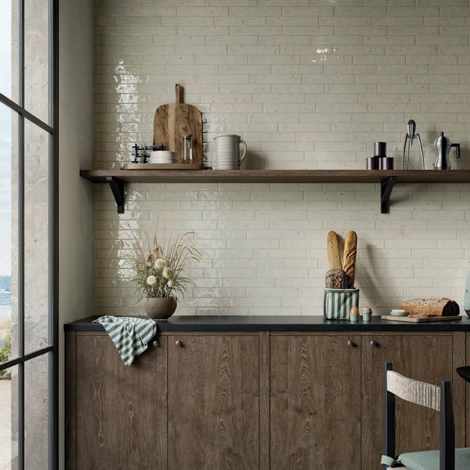 Modern kitchen with wooden cabinets, a black countertop, bread and kitchen utensils, a glass jar with flowers, and a striped towel. The space is beautifully finished with Via Del Sale - White tile backsplash in light beige subway style and open shelving displaying decor items.