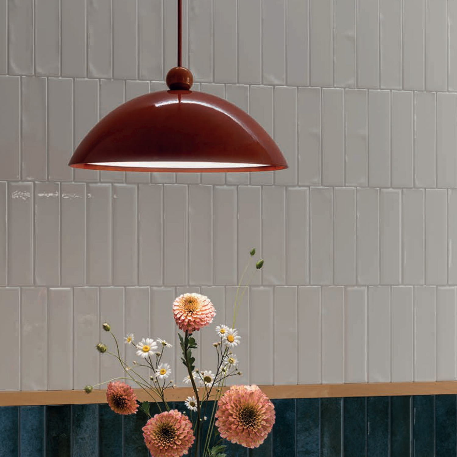 A red pendant lamp hangs over a vase with pink and white flowers, set against a wall featuring Vibe - Artic Snow glossy rectangular white tiles with a dark blue-green tiled lower section.