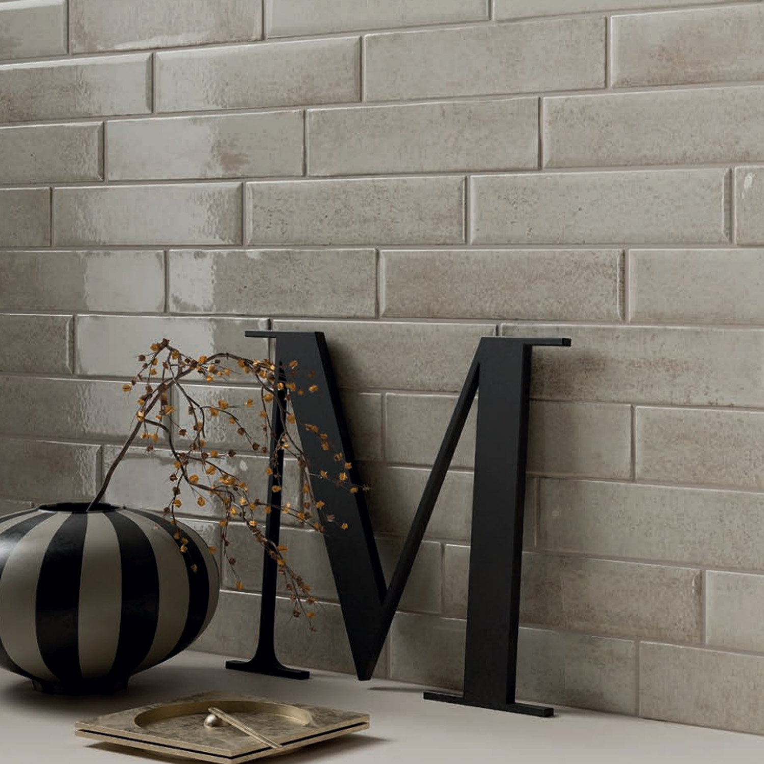 A black striped vase with a branch, a large black letter M, and a small square tray sit in front of a glossy Vibe - Bone tile wall.