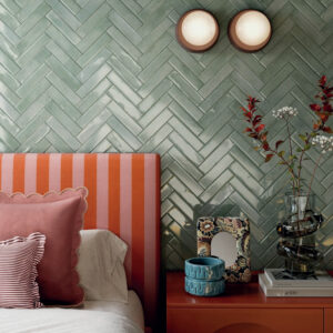 A bed with red and pink pillows sits next to an orange nightstand holding books, a vase of flowers, a blue dish, and a photo frame. The wall is adorned with Vibe - Mint Leaf green herringbone tiles and features two round lights.