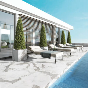 Step into a modern poolside patio featuring Bernini Classico marble flooring, complemented by potted topiary trees and several lounge chairs arranged to face the pool. The space boasts large glass windows and sleek architectural lines, all set beneath a clear blue sky.