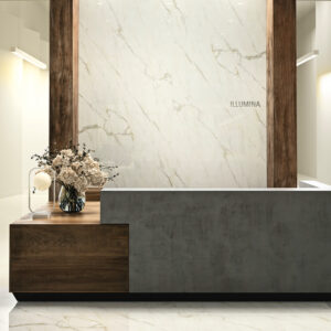 A modern reception desk featuring a concrete and wood finish stands before an elegant marble wall background of Oro Calacatta, engraved with the word ILLUMINA. A glass vase with a flower arrangement adds a touch of freshness to the clean, minimalistic setting.
